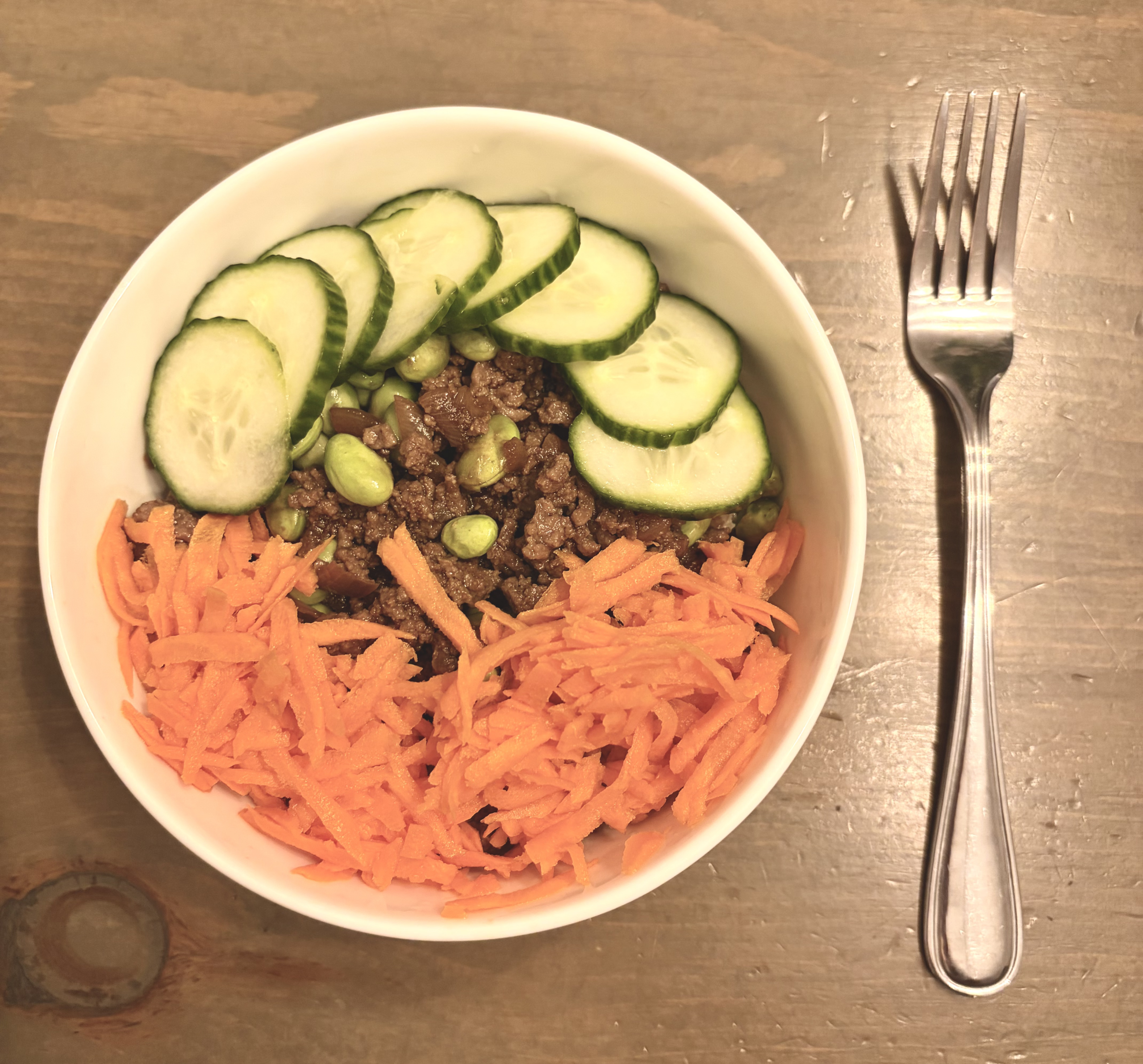 White bowl with ground beef, shredded carrots, cucumber slices, and edamame on a wooden table; a fork nearby.