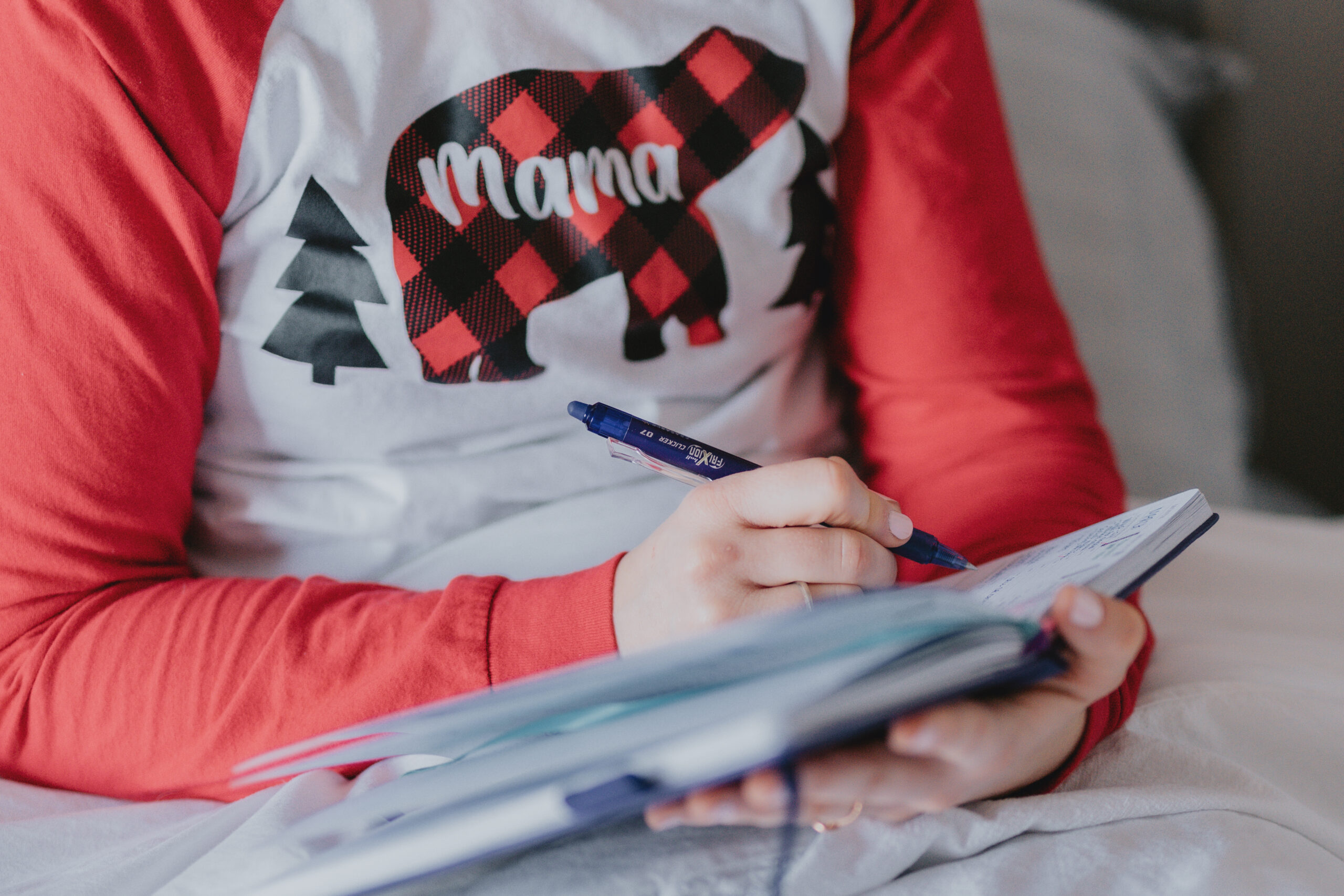Child in a red long-sleeve shirt writes in an open notebook with a blue pen on a light surface.