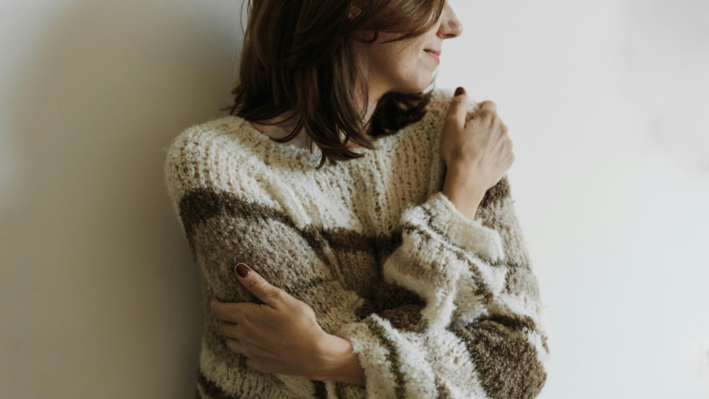 Woman in a chunky, striped knit sweater hugs herself while facing a light wall, seen in profile.