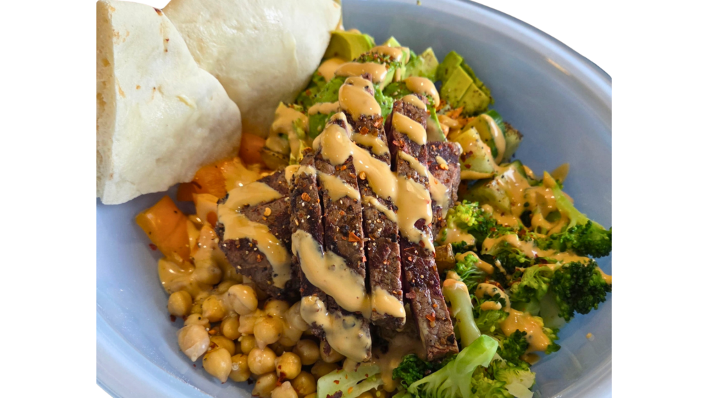 Sliced beef steak with a creamy sauce over broccoli and chickpeas on a blue plate with flatbread nearby.