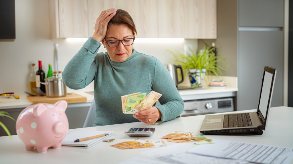 Woman in a teal top sits at a kitchen counter, holding money and touching her head in a worried gesture; a pink piggy bank, calculator, and scattered bills are visible on the table.