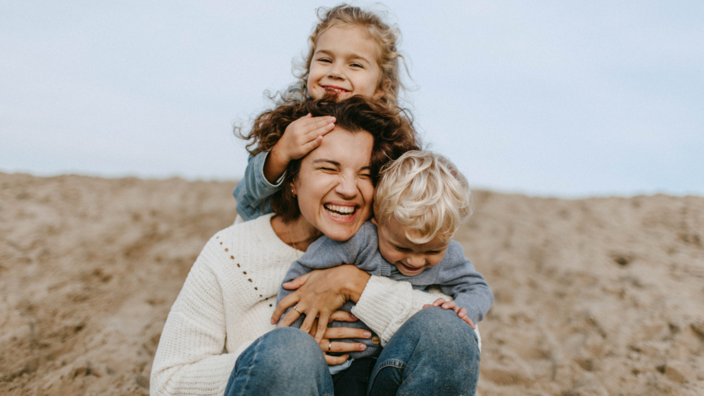 Mother laughing on a sandy beach, carrying her two children: a toddler on her shoulders and a baby in her arms.
