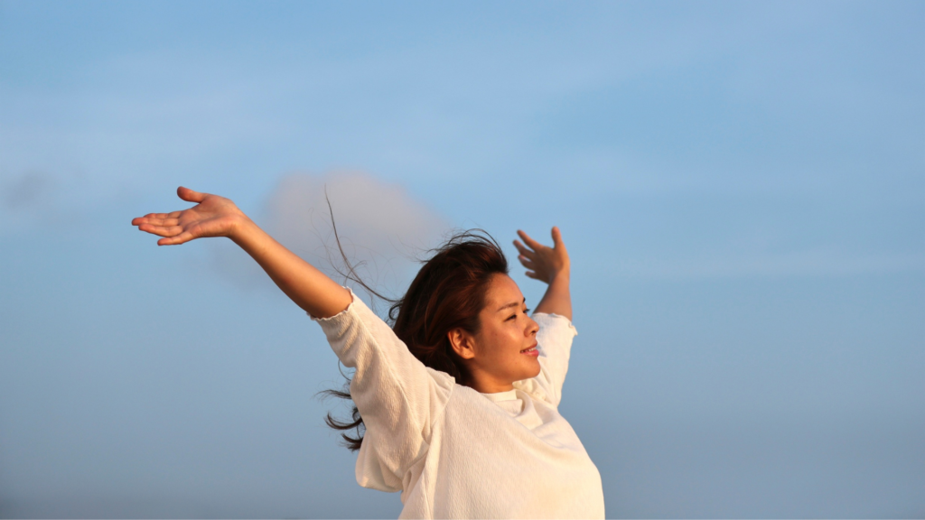 Woman with arms outstretched against a clear blue sky, smiling softly.