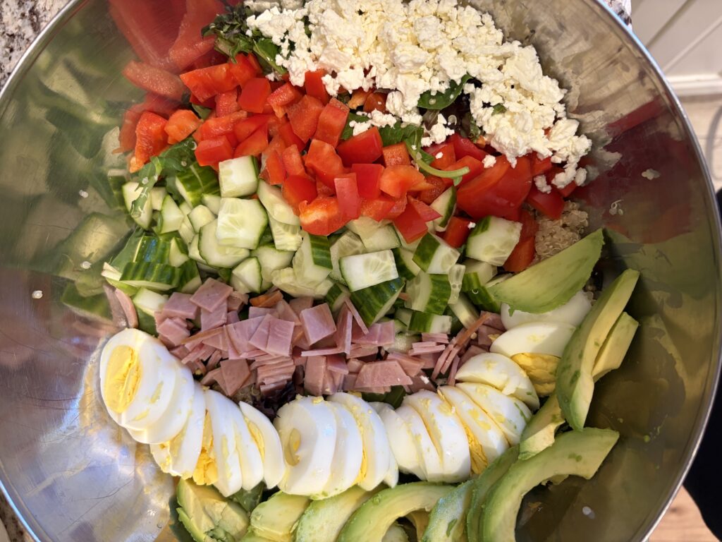Salad in a metal bowl with diced tomatoes, cucumbers, avocado slices, hard-boiled eggs, ham, feta, and greens.