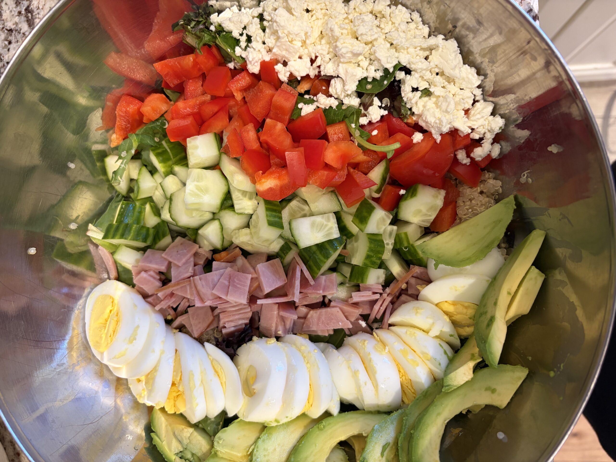 Salad in a metal bowl with diced tomatoes, cucumbers, avocado slices, hard-boiled eggs, ham, feta, and greens.