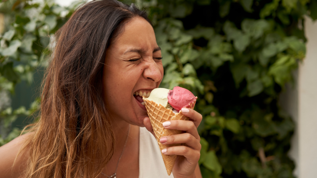 Woman taking a bite of a two-scoop ice cream cone outdoors (vanilla and strawberry).