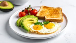 Breakfast plate with avocado slices, cherry tomatoes, toast, and two sunny-side-up eggs on a white plate.