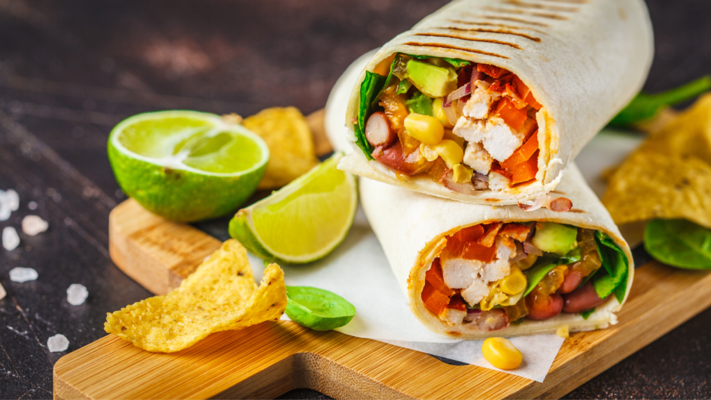 Two grilled wraps cut in half, filled with chicken, corn, beans, avocado, peppers and greens, on a wooden board with lime wedges and tortilla chips.