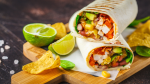 Two grilled wraps cut in half, filled with chicken, corn, beans, avocado, peppers and greens, on a wooden board with lime wedges and tortilla chips.