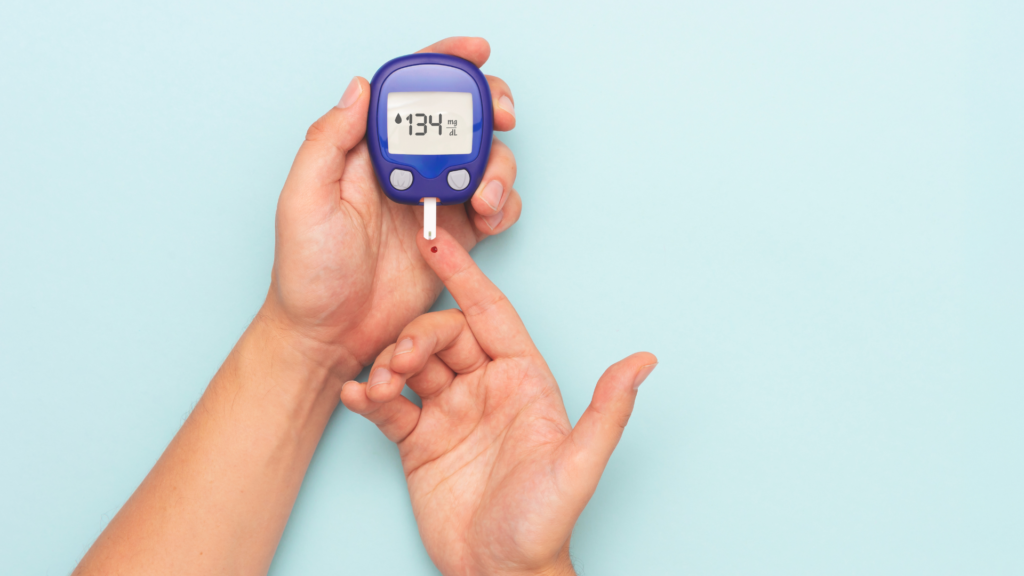 Hand holding a blue glucometer while a finger with a blood drop is being tested for glucose.