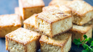 Golden-brown cornbread cubes with a crumbly interior, lightly dusted and piled on a dark surface, with parsley garnish in the corner.