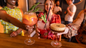Friends raise colorful cocktails for a toast at a bar, smiles in the background.