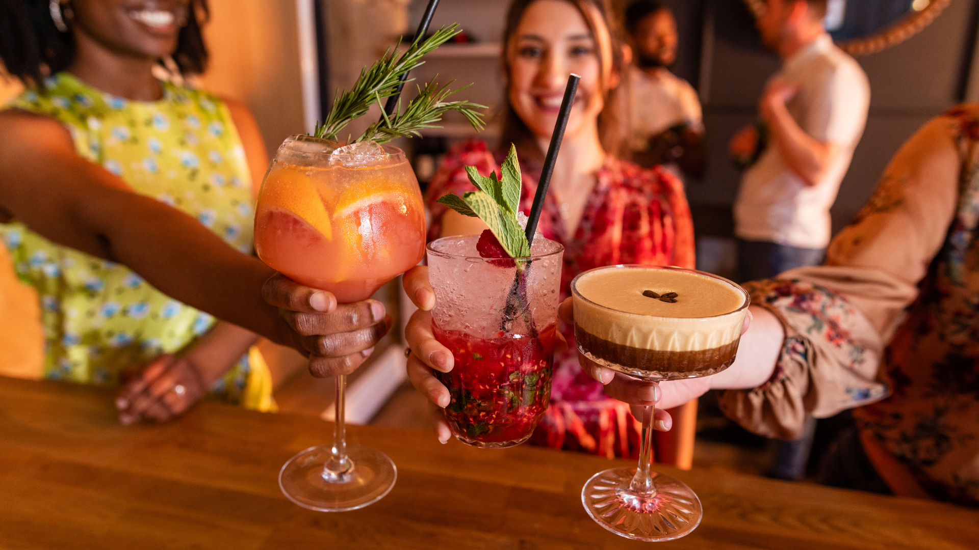 Friends raise colorful cocktails for a toast at a bar, smiles in the background.