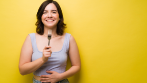 Smiling woman holds a fork in front of a bright yellow wall, suggesting ready to eat or dining.