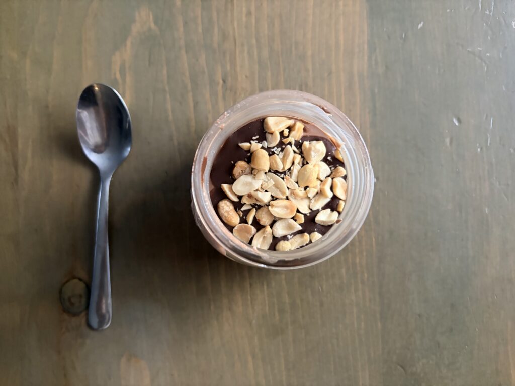 Top-down view of a chocolate dessert in a glass jar, topped with crushed peanuts, with a spoon to the left on a wooden table.