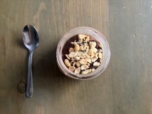 Top-down view of a chocolate dessert in a glass jar, topped with crushed peanuts, with a spoon to the left on a wooden table.